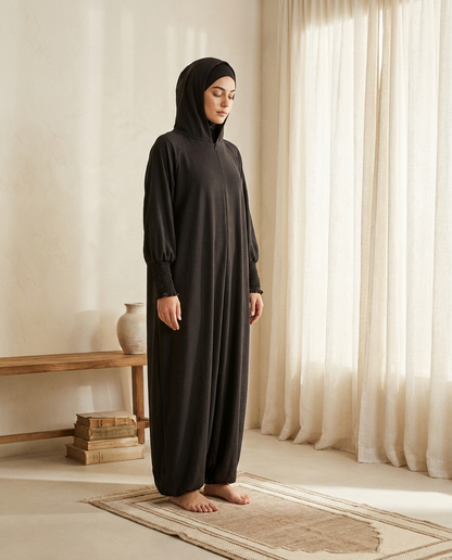 Woman wearing black hooded prayer jilbab standing on a prayer mat in soft morning light